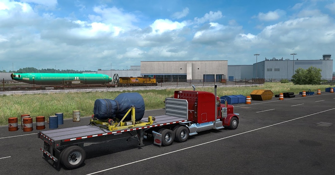 Ein Lastwagen mit Tieflader transportiert abgedeckte Flugzeugkomponenten in der Nähe einer Fertigungsanlage. Im Hintergrund ist ein Flugzeugrumpf auf einem Schienenwagen zu sehen, was auf eine komplexe Logistikoperation hindeutet.